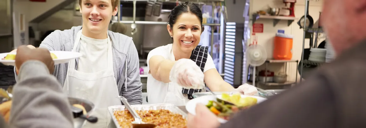 Two volunteers serving hot meals to people at a community kitchen counter