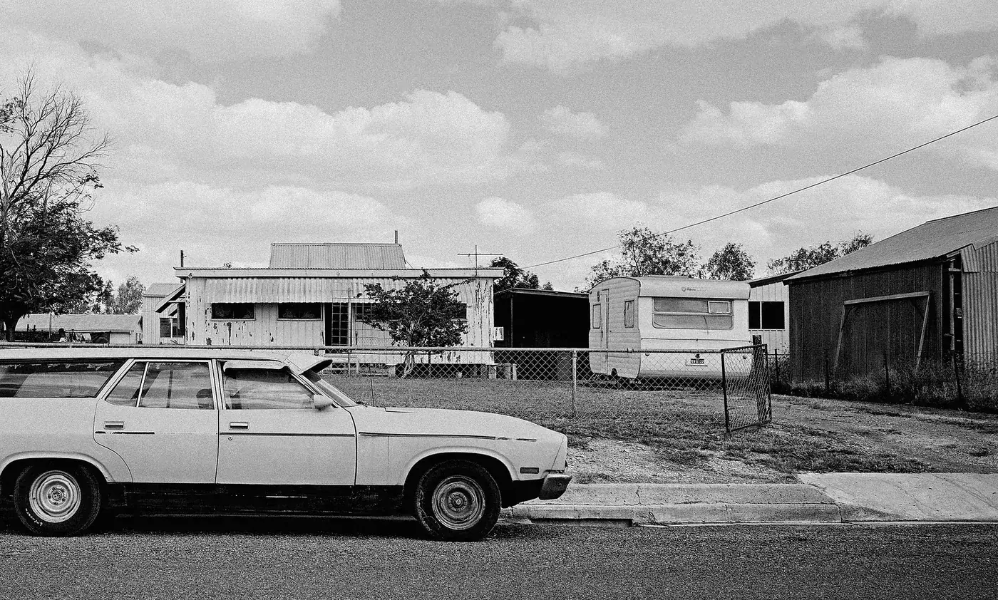 A quiet rural street with a weathered house, a car, and a trailer